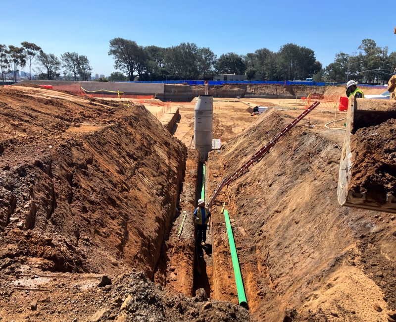 Worker installs green pipes in trench; equipment and barriers nearby.