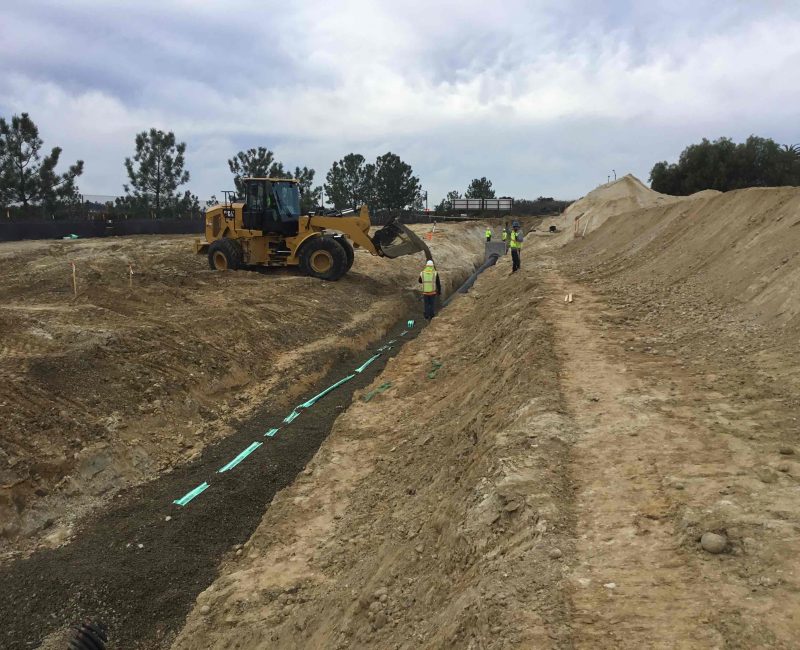 Workers watch a yellow front loader fill a green-lined trench.
