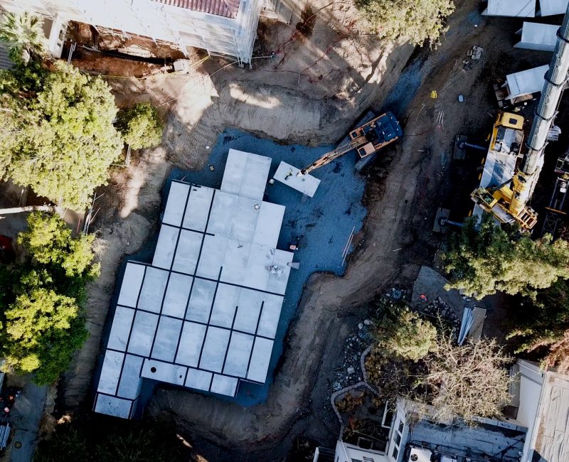 Aerial shot of construction site, foundation, vehicles, trees visible.