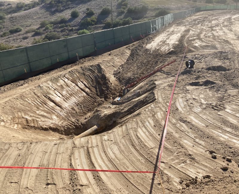 Deep construction pit, exposed pipes, red tape, green fence, hills behind.