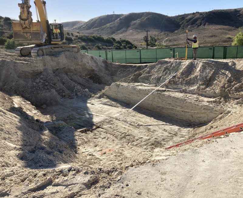 Excavator and worker measure pit at construction site; hills behind.
