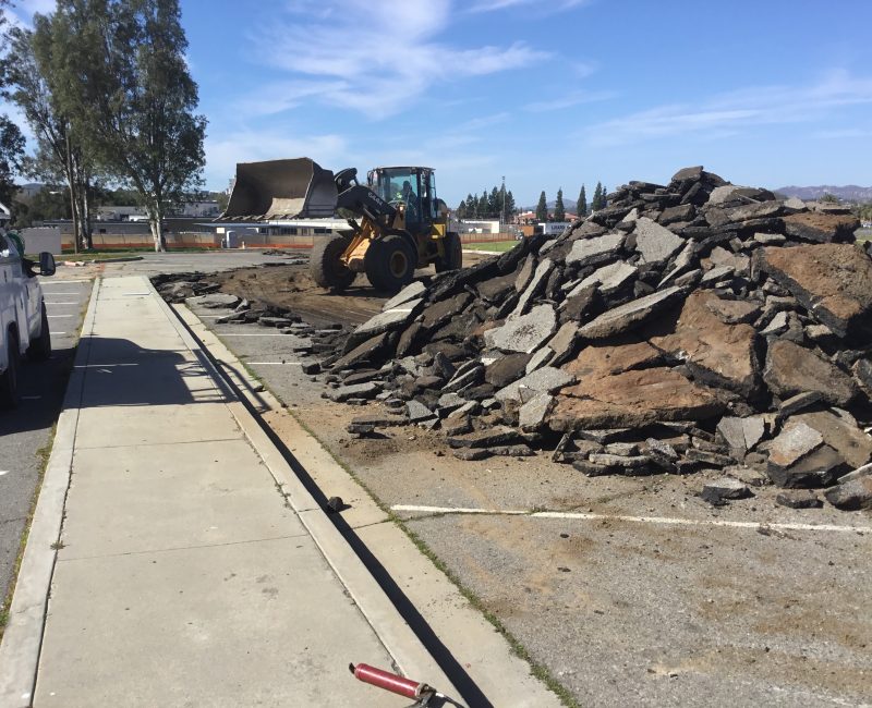 Bulldozer clears asphalt debris by sidewalk; trees in background.