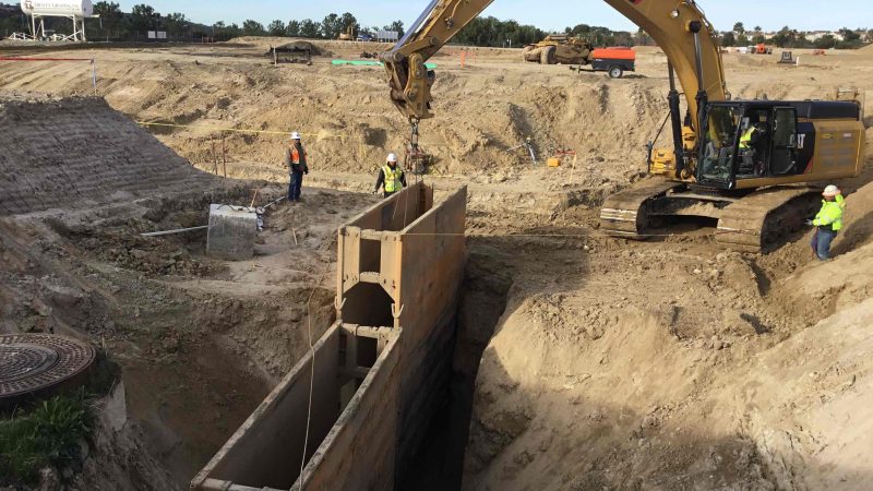 Workers and excavator by trench with steel supports, clear sky.
