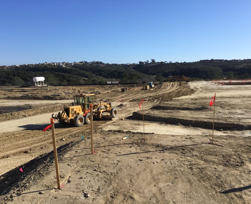 Construction site with grader, dirt roads, flags, hills, blue sky.