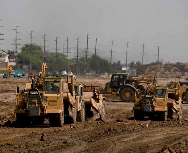 Construction vehicles work on a dusty site with buildings behind.