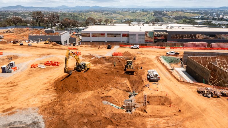 Construction site with machines, materials, half-built structures, and hills.