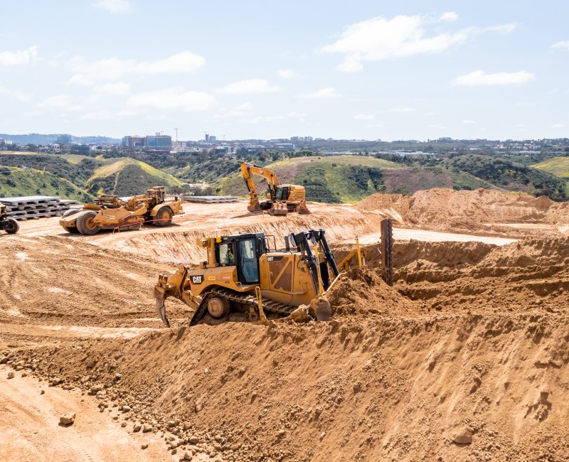 Bulldozers and excavators move dirt at sunny hilltop site.