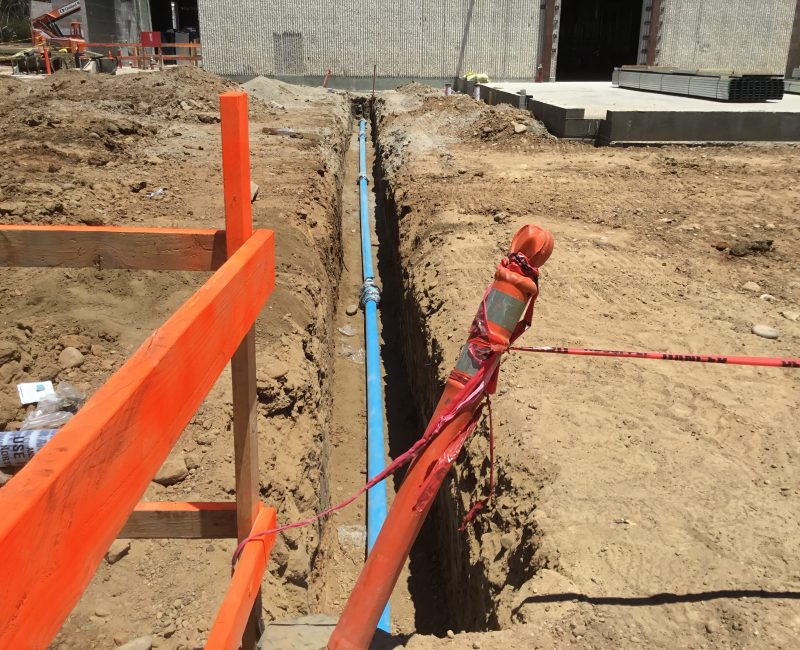 Blue pipe in deep trench, dirt, orange barriers, building behind.