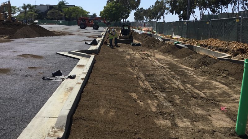 Workers prepare dirt by road; equipment, fences, and trees visible.