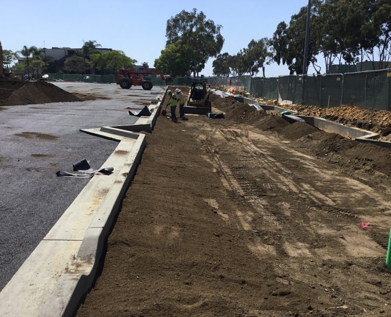 Workers prepare dirt by road; equipment, fences, and trees visible.