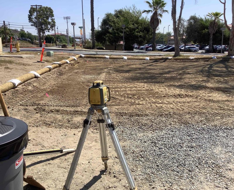 Yellow tripod on gravel at construction site, trees and cars behind.