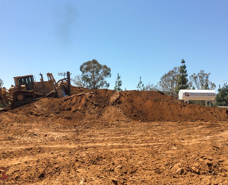 Bulldozer on a dirt mound, Trinity Grading sign, trees behind.
