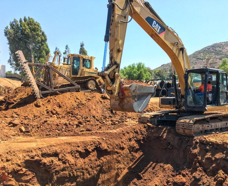 Yellow excavator digs dirt; bulldozer nearby, trees and hills behind.