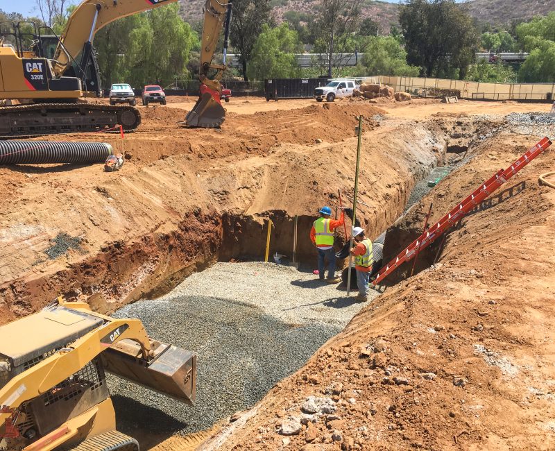 Three workers in vests and helmets stand in a deep trench.