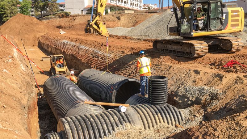 Workers install black drainage pipes in trench with equipment.