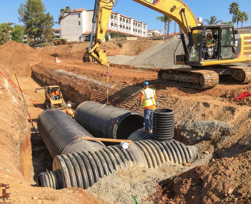 Workers install black drainage pipes in trench with equipment.