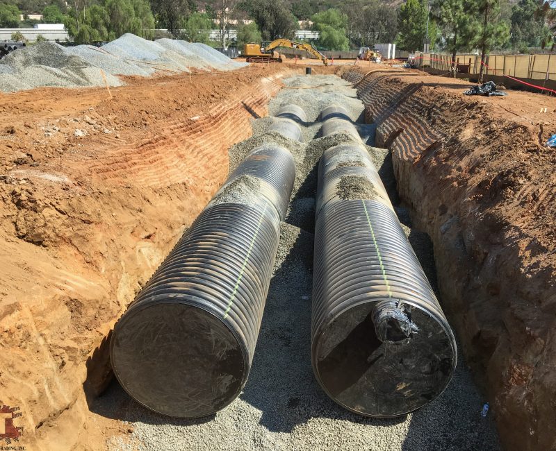 Two large black pipes lie side by side in a trench.