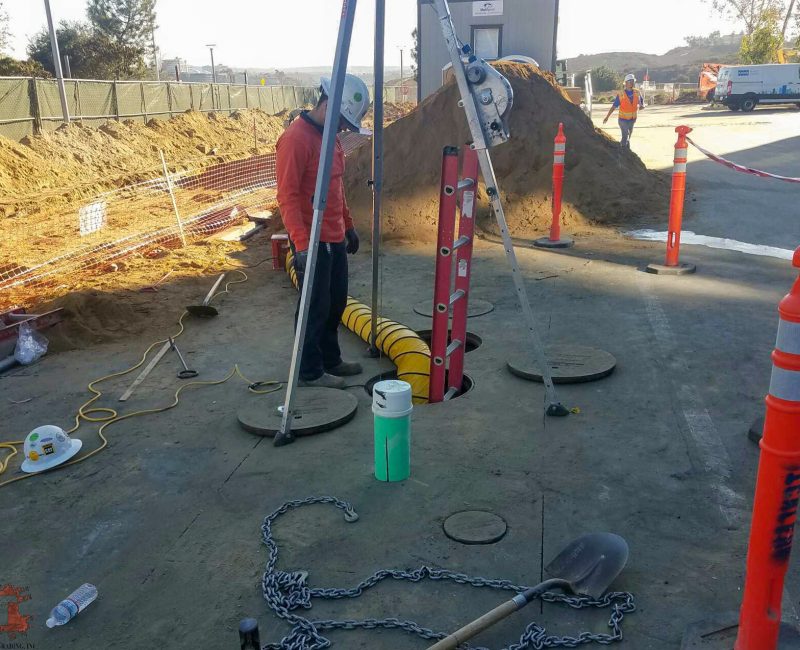 Worker in helmet by open manhole, tools and cones nearby.