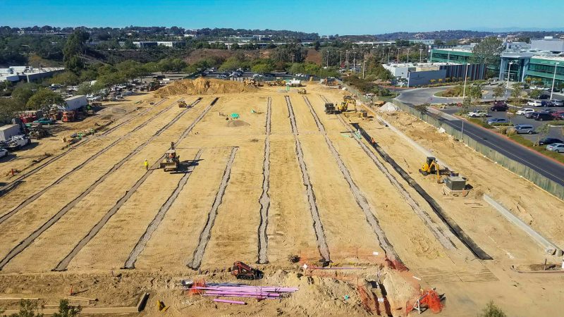 Construction site with bulldozers, trenches, nearby buildings, clear sky.