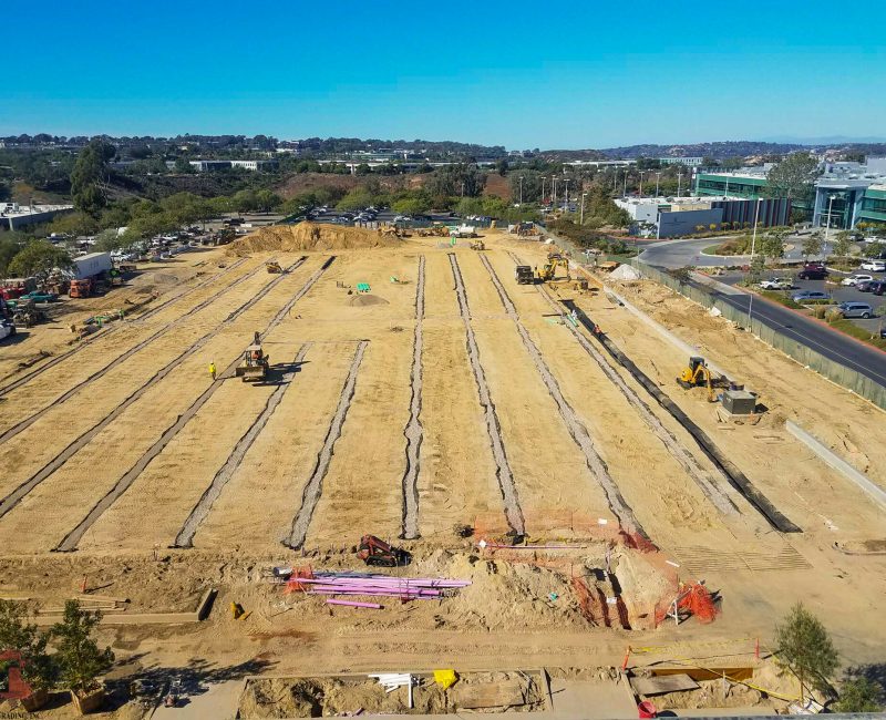 Construction site with bulldozers, trenches, nearby buildings, clear sky.