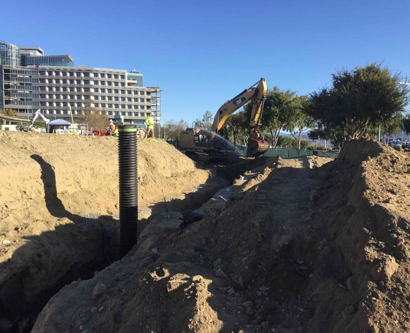 Excavator digging trench near upright drainage pipe at construction site.