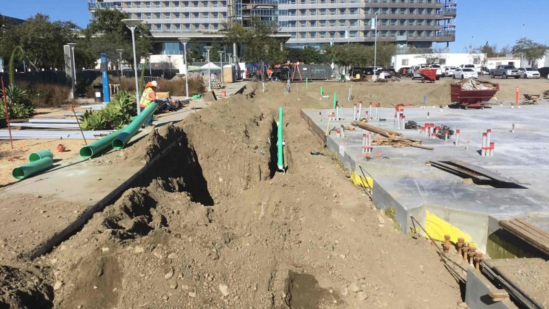 Construction site with trench, green pipes, workers, building, and vehicles.