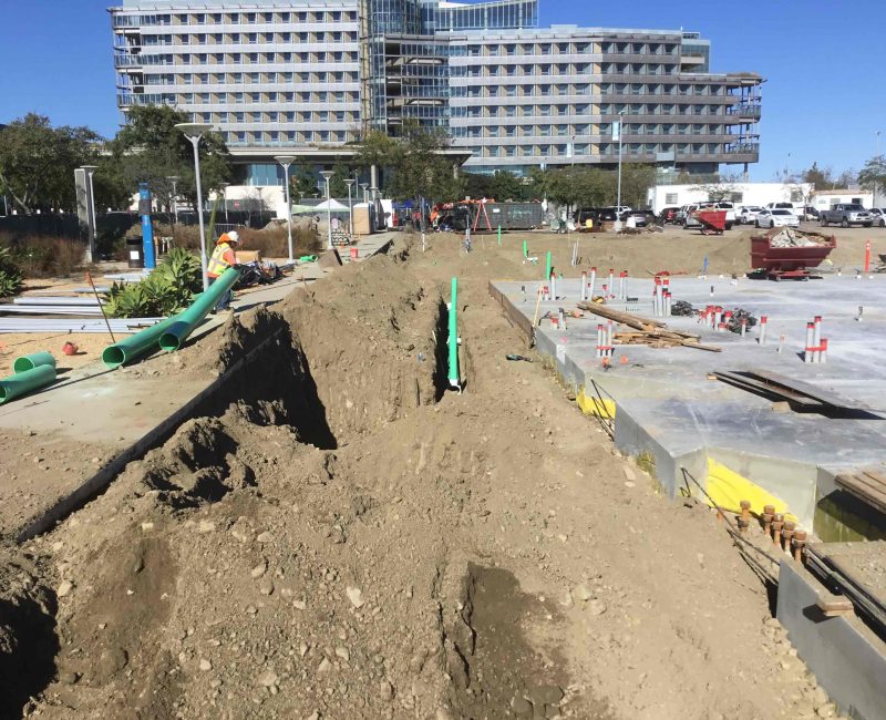 Construction site with trench, green pipes, workers, building, and vehicles.