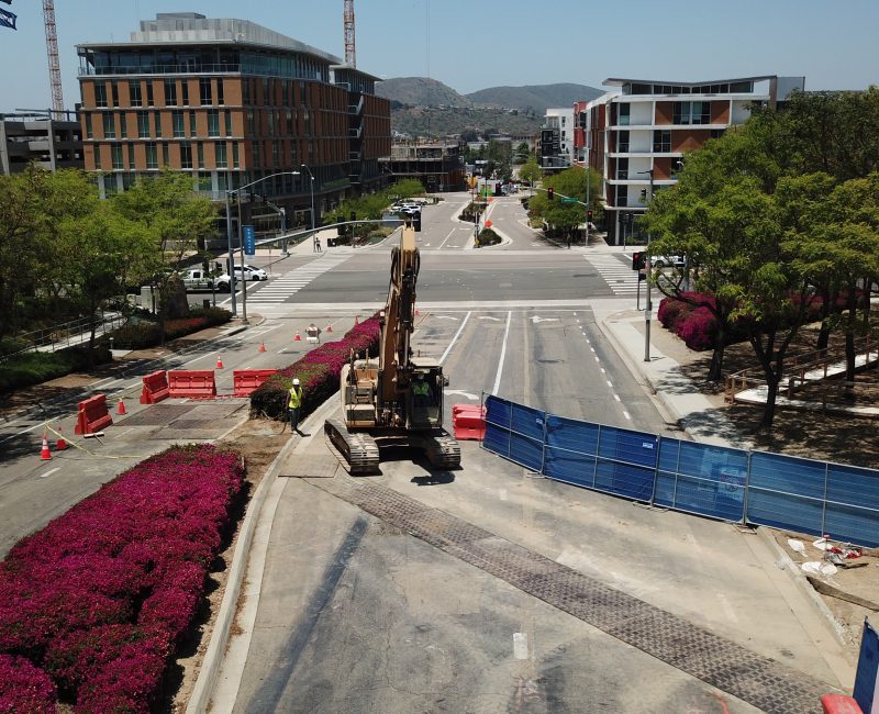 Excavator and barriers block city street; buildings, bushes nearby.