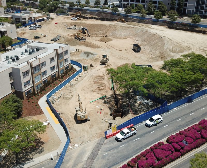 Construction site by residences, blue fence, white vans, pink bushes.