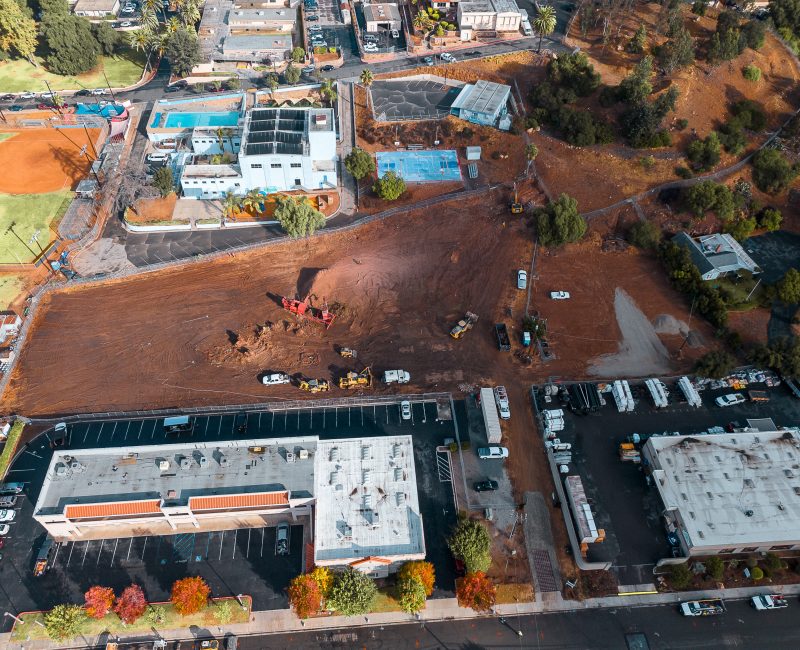 Aerial view of construction site, machines, parking lots, buildings nearby.