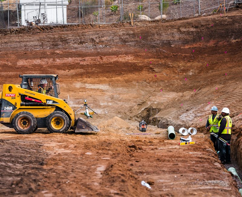 Bulldozer and workers with pipes at site, pink markers visible.