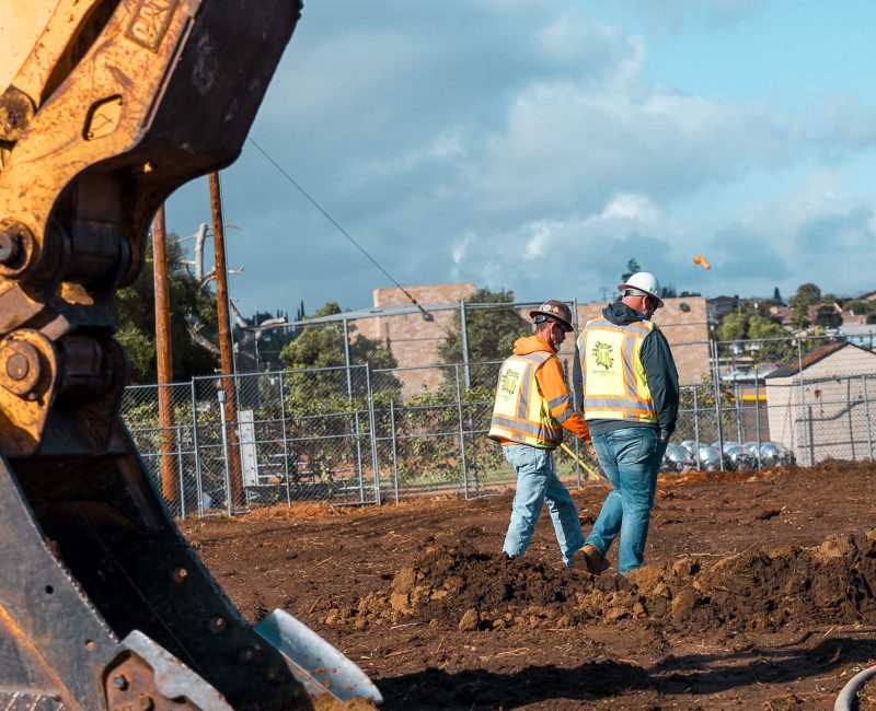 Two workers in safety gear stand by a large excavator outside.