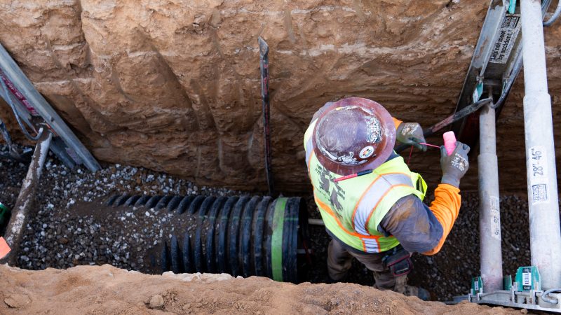 Worker in hard hat stands in trench with pipes and equipment.