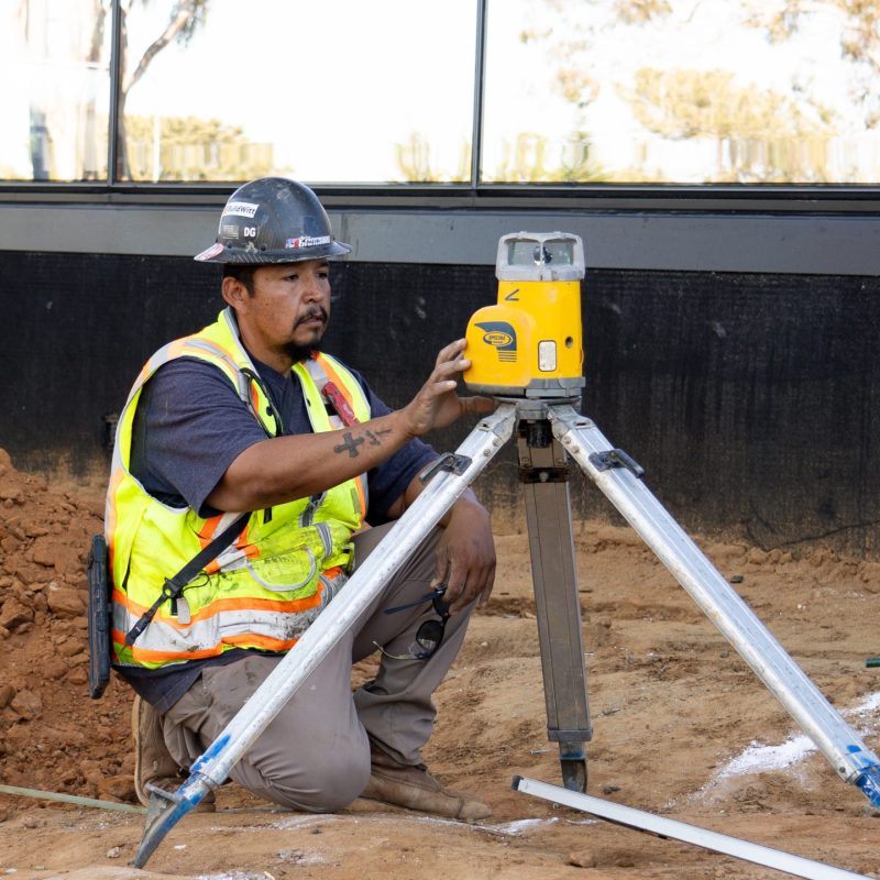 Worker in vest uses yellow surveying tool at construction site.