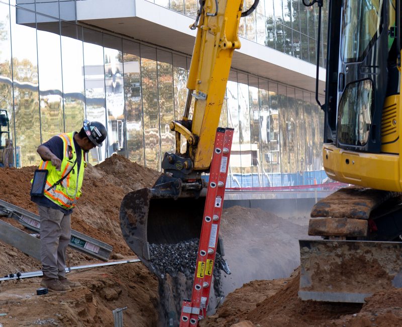 Worker by excavator pouring dirt into trench by glass building.