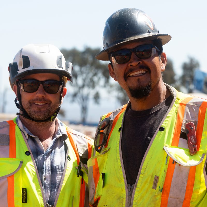 Two smiling construction workers in safety gear at outdoor site.