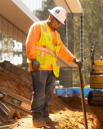 Worker in trench with tool; excavator and sunlight in background.