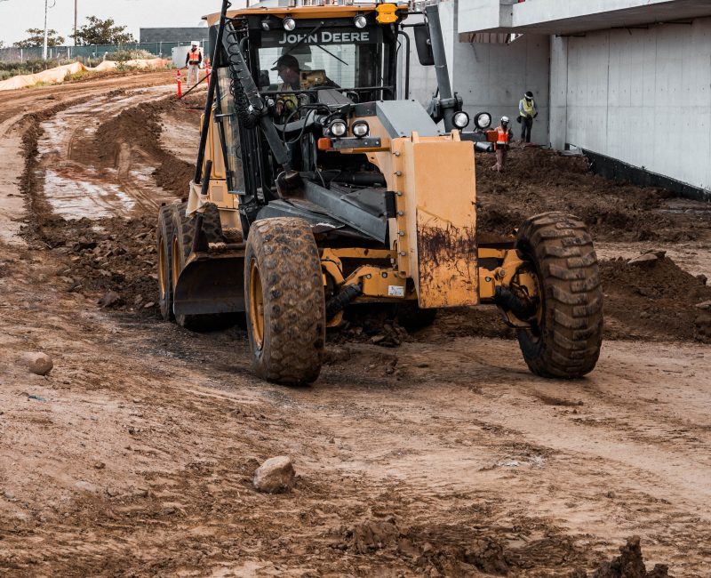 Worker uses yellow grader near building site; others in background.