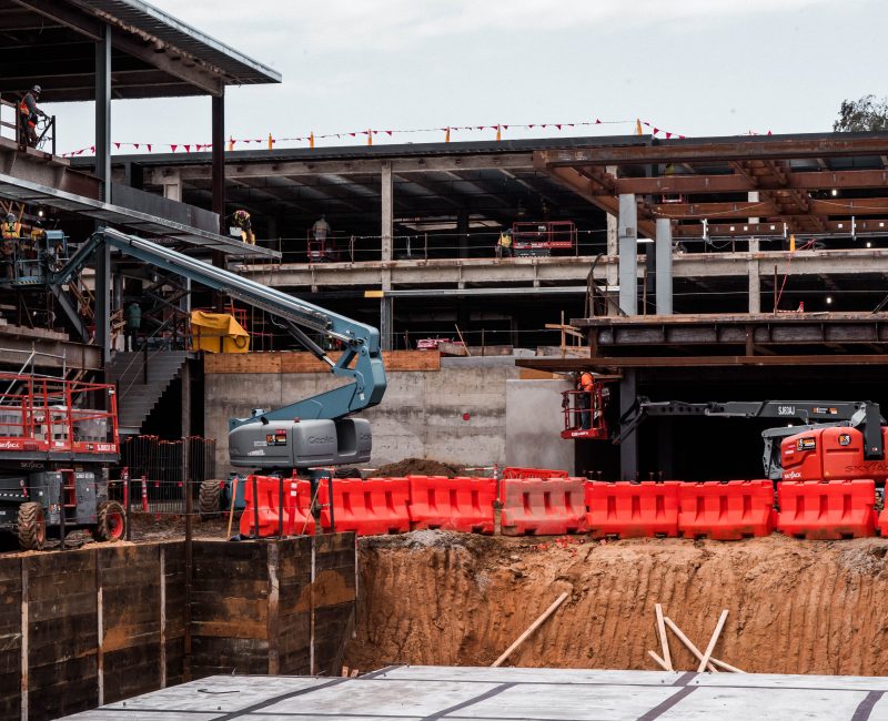 Steel framework, workers, red barriers, excavators, and a construction pit.