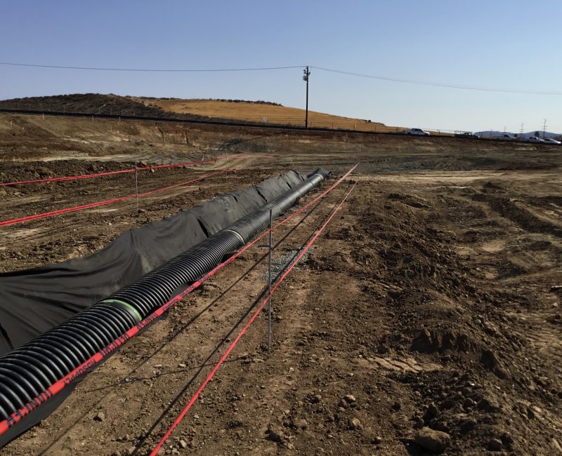 Black drainage pipes in fabric-lined trench, red tape nearby.