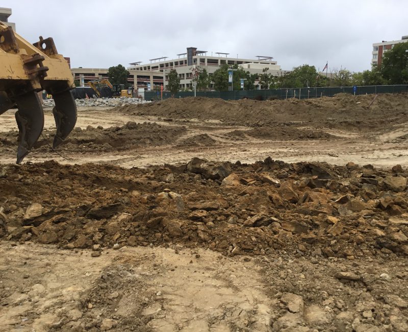 Construction site with machinery, uneven dirt, rocks, and buildings.