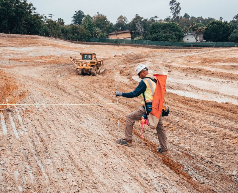 Worker measuring distance at dirt site; bulldozer in background.