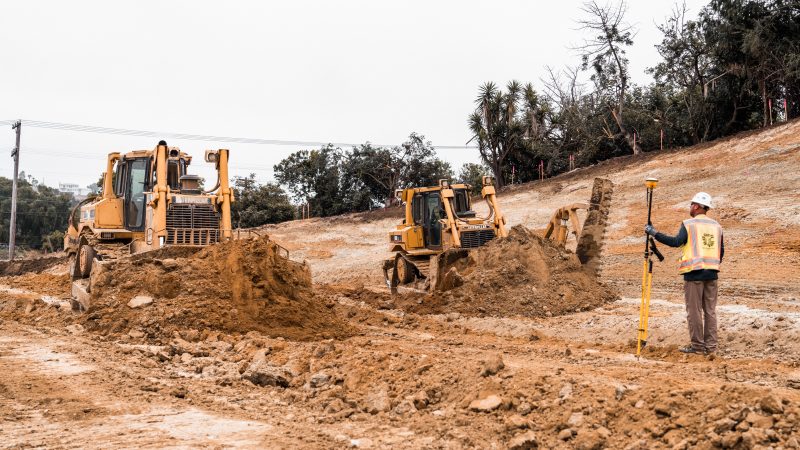 Bulldozers move dirt; worker surveys site; trees, wires behind.