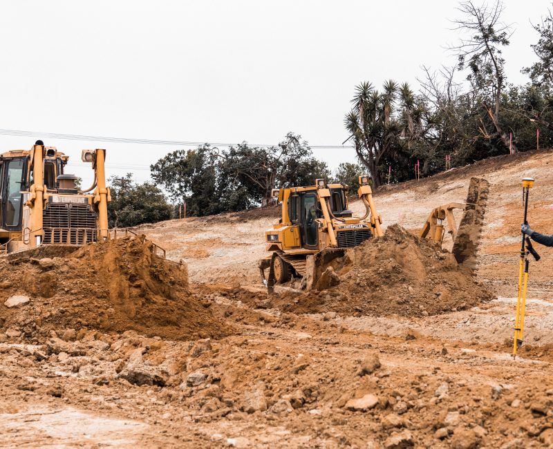 Bulldozers move dirt; worker surveys site; trees, wires behind.