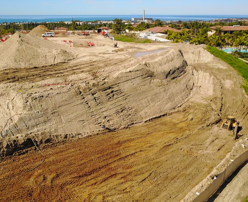 Construction site with dirt mounds, equipment, houses, ocean in distance.