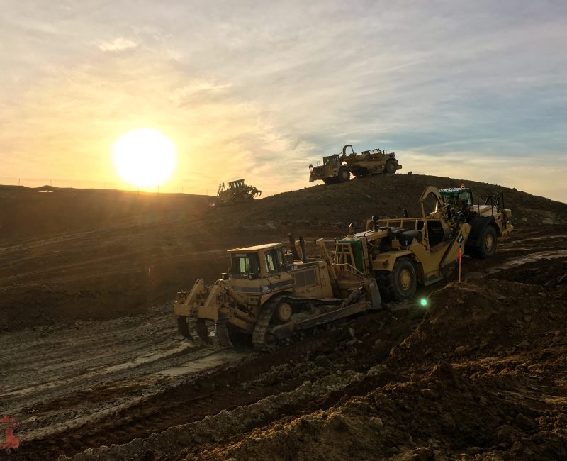 Bulldozers and loaders on dirt hill at sunrise, tire tracks visible.