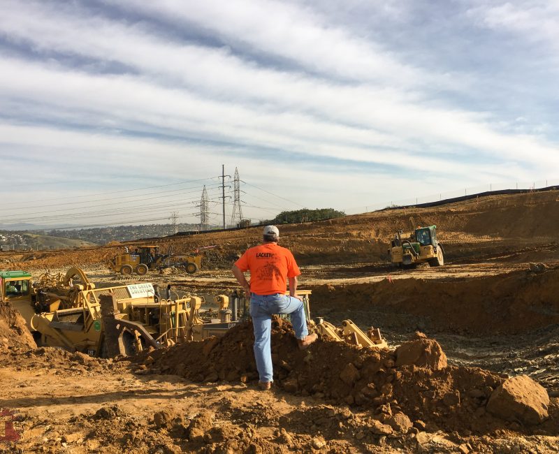 Man in orange shirt watches construction vehicles on a dirt mound.