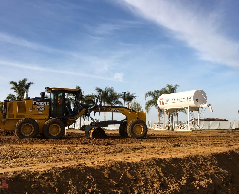 Yellow John Deere grader at construction site, palm trees, blue sky.