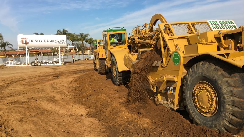 Yellow construction vehicles move dirt; Trinity Grading sign, palm trees.