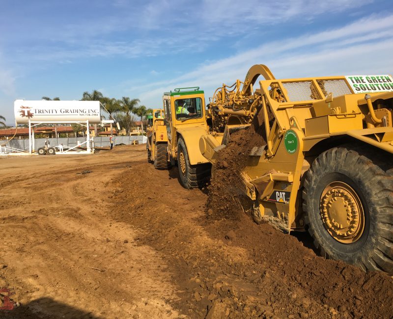 Yellow construction vehicles move dirt; Trinity Grading sign, palm trees.